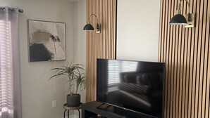 Un salon moderne doté d’un téléviseur à écran plat, d’un mur recouvert de panneaux en bois, de deux lampes suspendues, d’une plante en pot et d’un tapis gris.