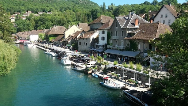 A riverside town with boats docked along the water, buildings with balconies, and a pedestrian walkway.