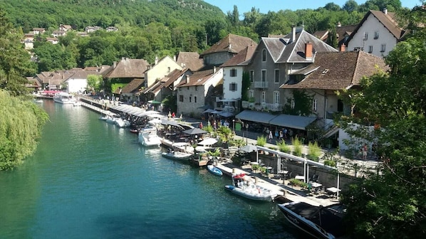 A riverside town with boats docked along the water, buildings with balconies, and a pedestrian walkway.