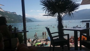 A beachside view from an outdoor seating area with tables and chairs, overlooking a crowded beach with people swimming and sunbathing.