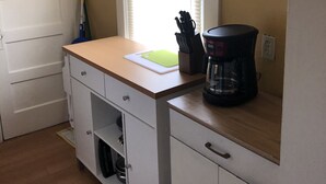Une cuisine de taille réduite, dotée d’un plan de travail blanc, d’une machine à café et d’une fenêtre avec des stores.