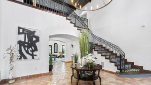Un hallo amplio con piso de baldosas, una escalera con barandillas decorativas y una mesa redonda con un jarrón de flores.