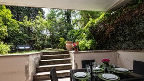 Eine Terrassensitzgruppe im Freien mit Tisch, umgeben von üppigem Grün und einer Steintreppe, die zu einer höheren Ebene führt.