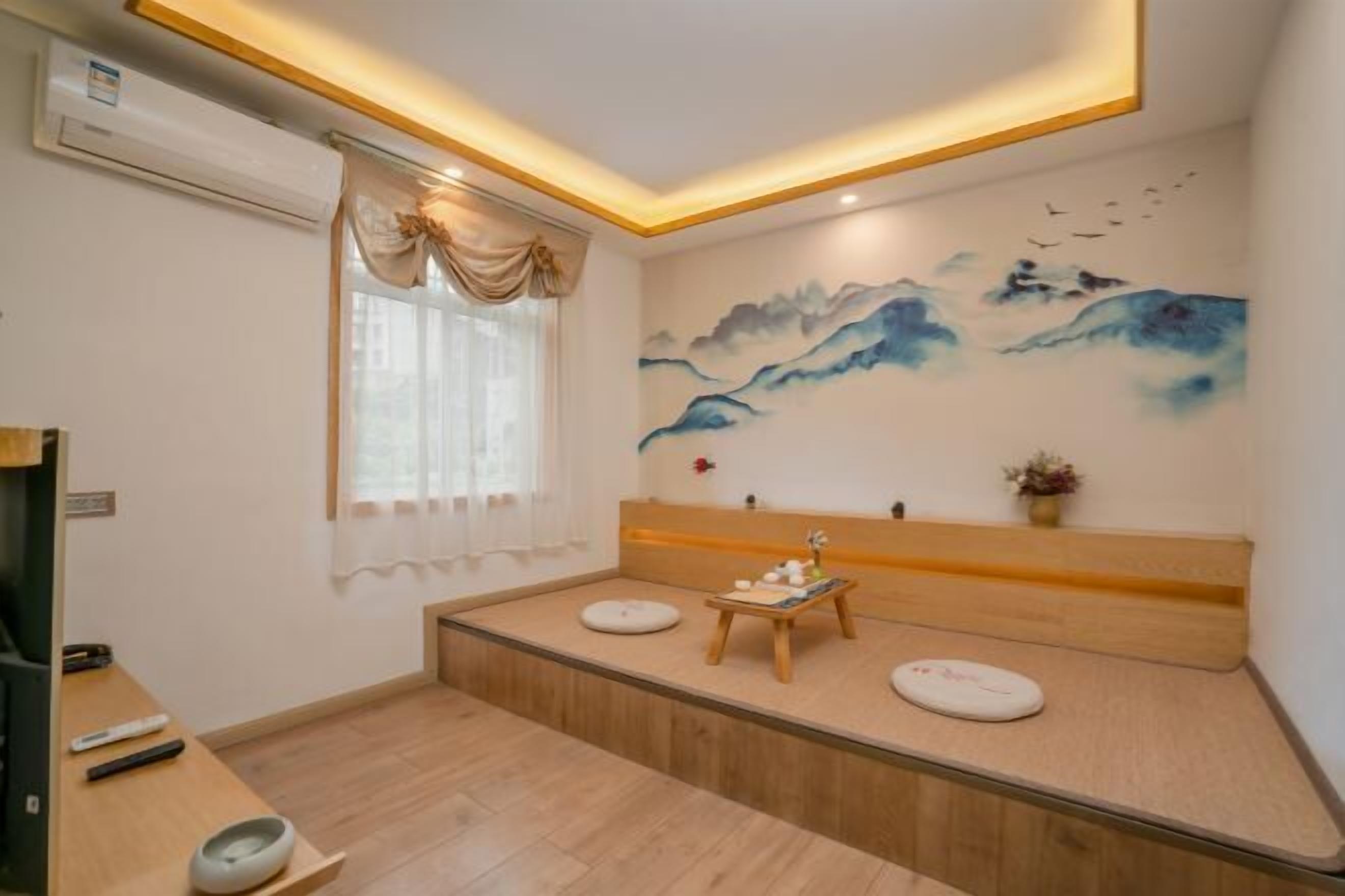 Quarto com um banco de madeira, ar condicionado fixado na parede, janela com cortinas e um quadro de montanhas.