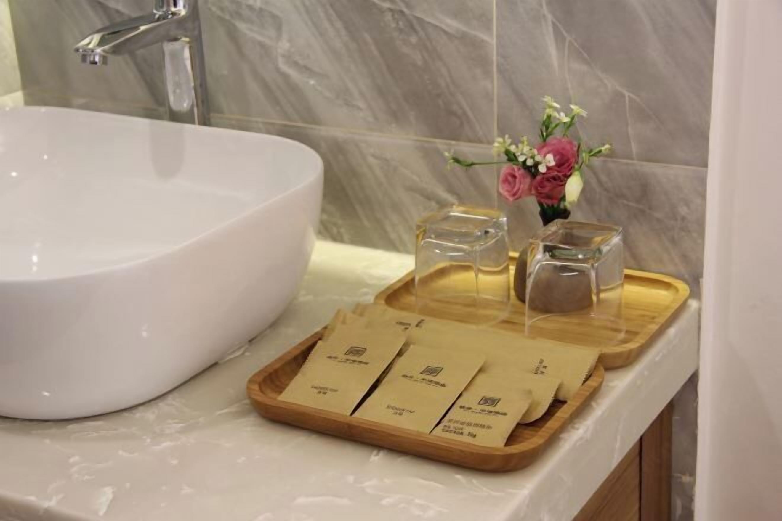 A bathroom with a white sink, a marble wall, a tray with toiletries, and a vase with flowers.