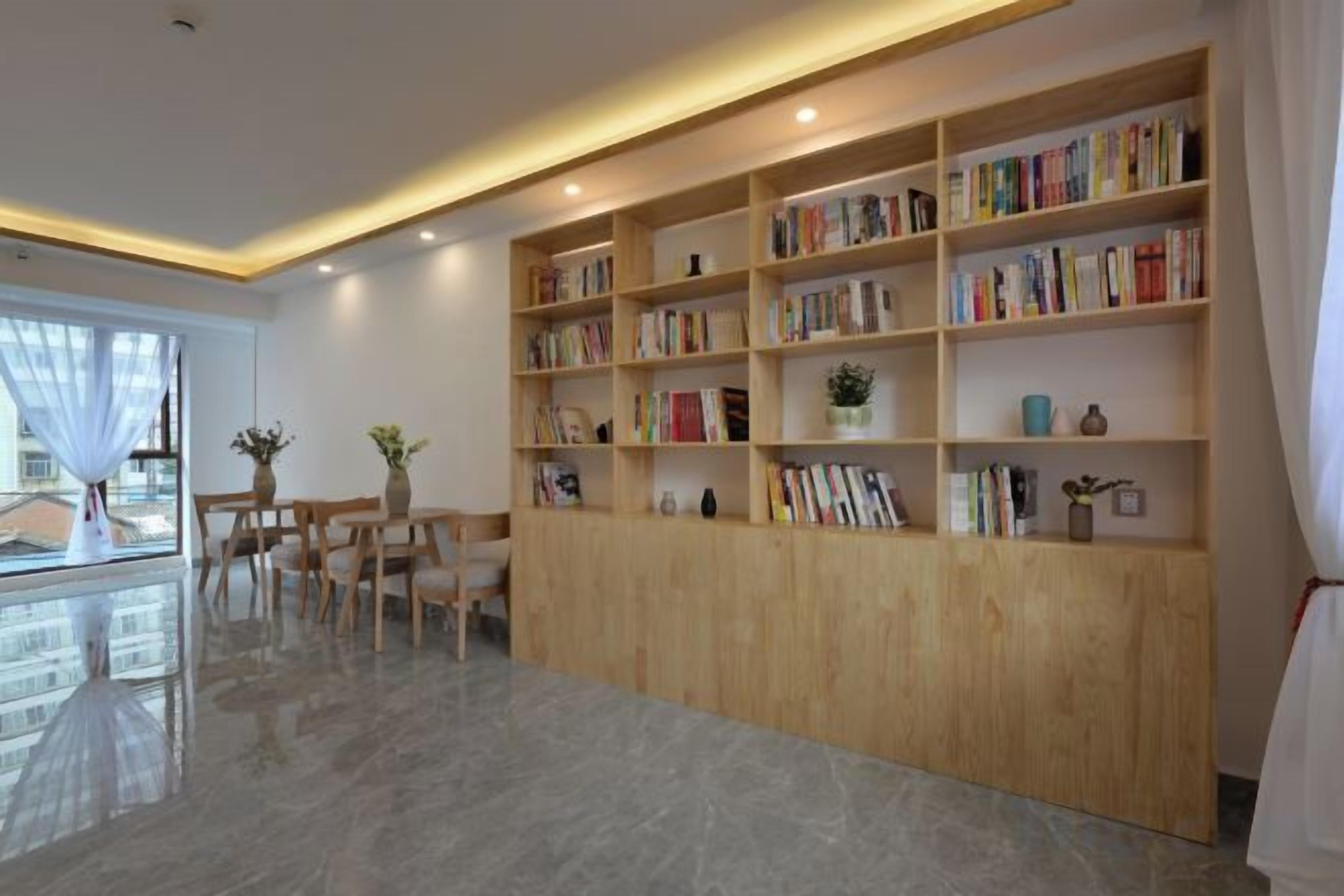 A modern interior with a wooden bookshelf filled with books, a dining area with chairs, and a large window providing natural light.