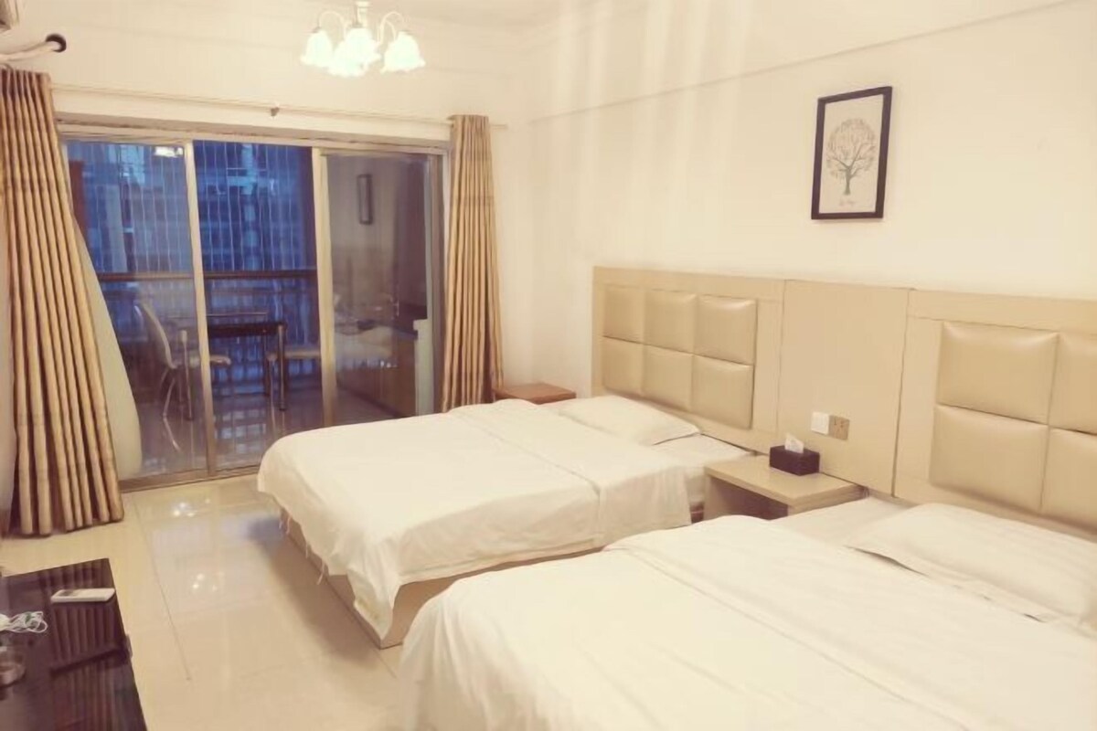 Habitación de hotel con dos camas, un ventanal grande con cortinas, una mesita de noche con lámpara y un cuadro enmarcado en la pared.