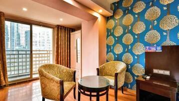 A room with two patterned armchairs, a small glass table, a fireplace, and a large window with curtains.