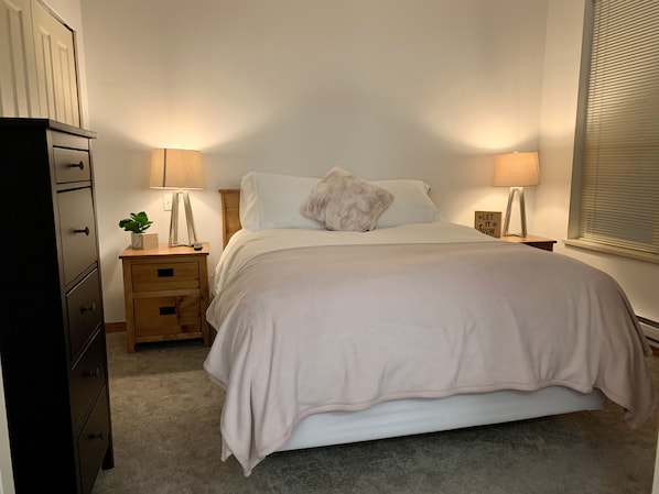 A bedroom with a bed, two bedside tables, a nightstand, a dresser, and a window with blinds.