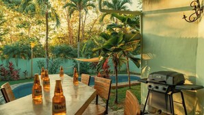 Un espacio de comedor al aire libre con una mesa preparada para cuatro personas, rodeada de sillas, una parrilla y botellas de cerveza sobre la mesa.