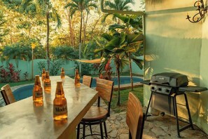 An outdoor dining area with a table set for four, surrounded by chairs, with a grill and bottles of beer on the table.