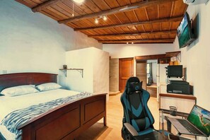 A bedroom with a wooden ceiling, a bed, a desk with a chair, a laptop, and a television mounted on the wall.