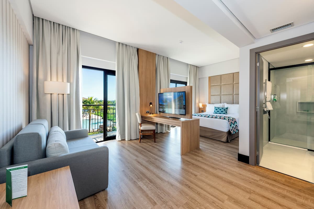 A modern hotel room with a sofa, a desk with a TV, a bed, and a glass shower area.