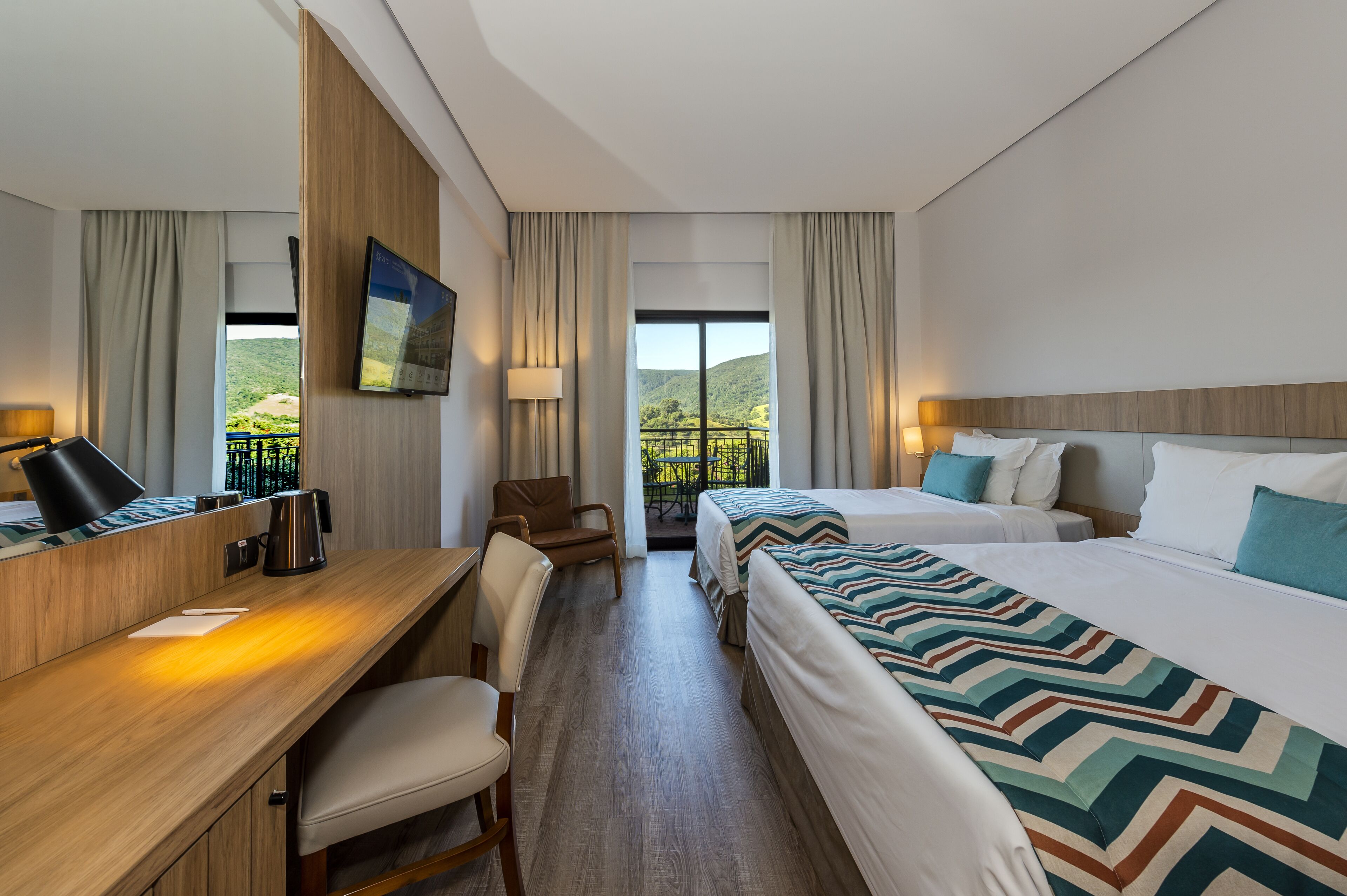 A hotel room with two beds, a desk, a chair, a TV, and a balcony with a view of greenery.