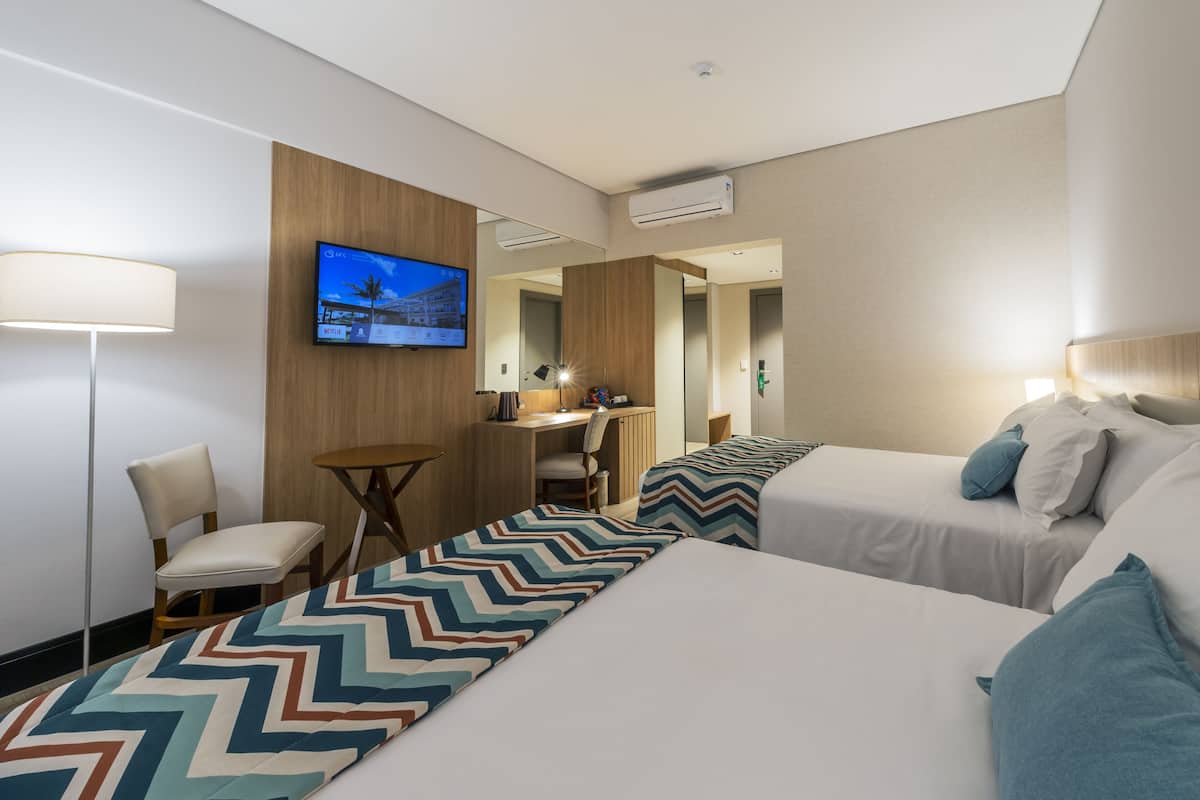 A hotel room with two beds, a desk, a chair, a TV mounted on the wall, and a mirror.