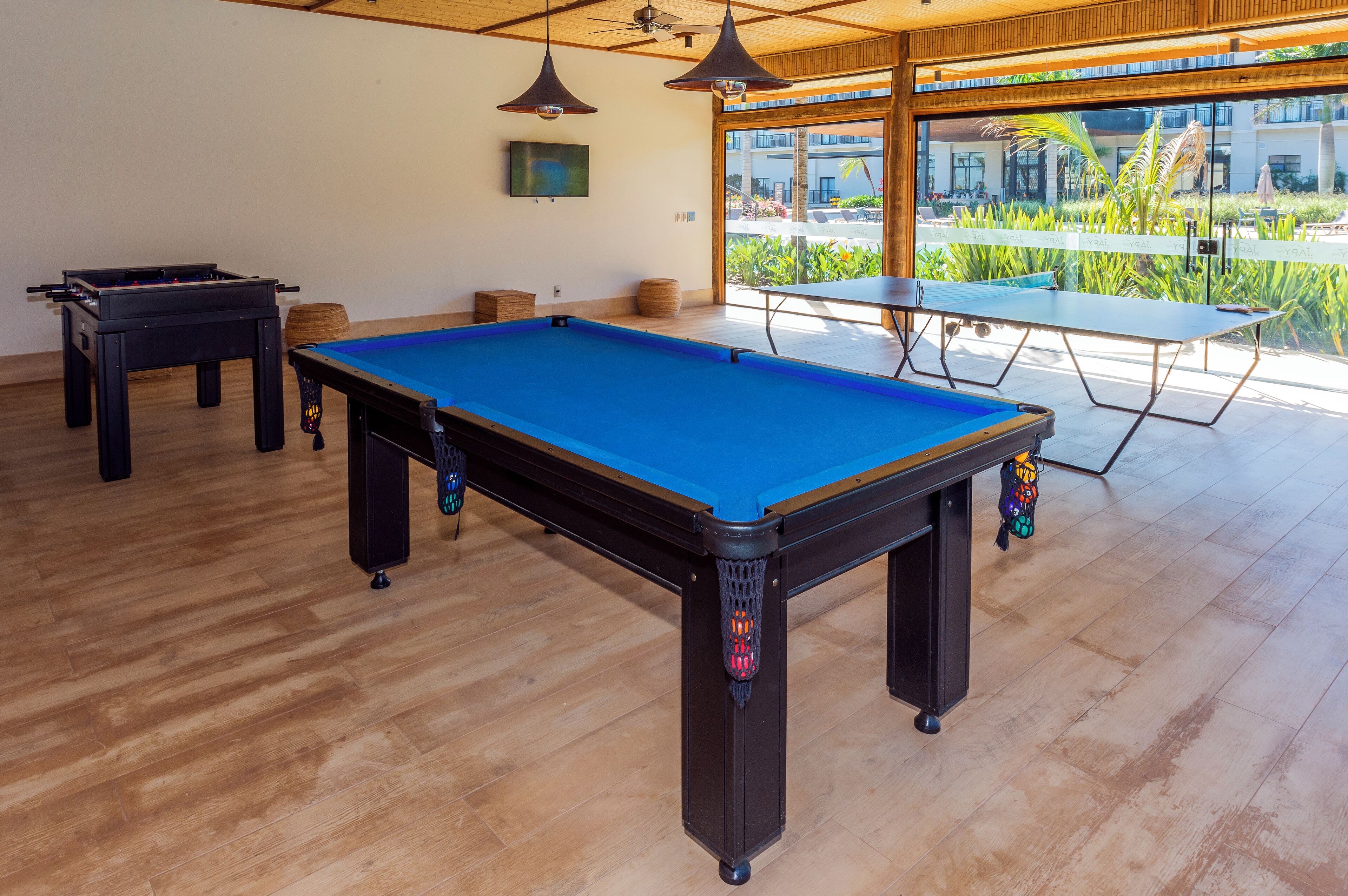 A pool table with a blue felt surface, a foosball table, and a ping pong table in a room with wooden flooring and large windows.