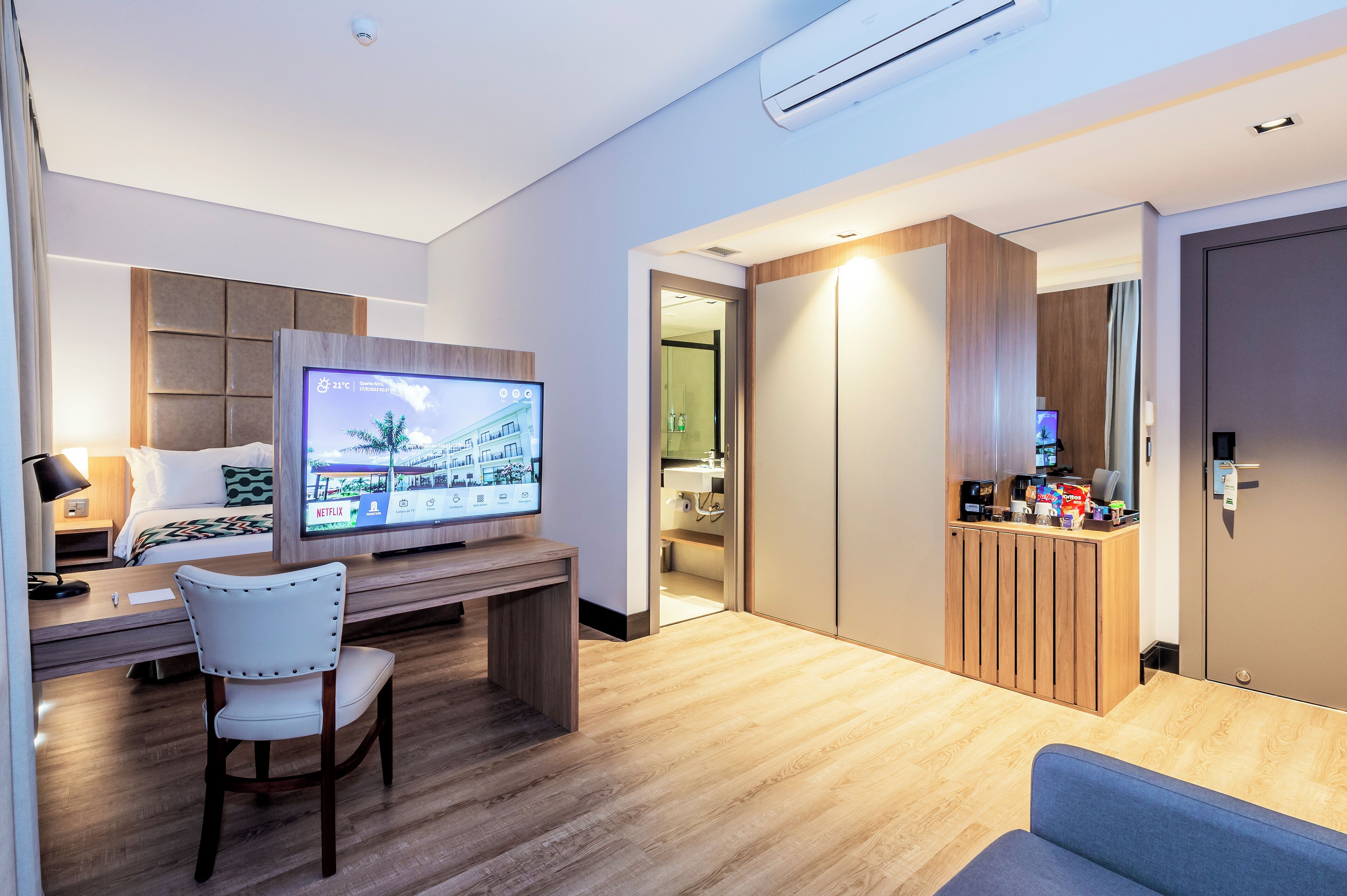 A modern hotel room with a bed, a desk with a TV, a chair, a sofa, and a minibar.
