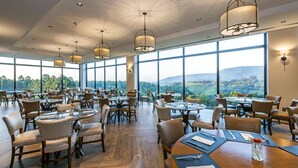 Un comedor amplio con varias mesas y sillas, ventanas grandes que ofrecen vistas al paisaje e iluminación colgante.