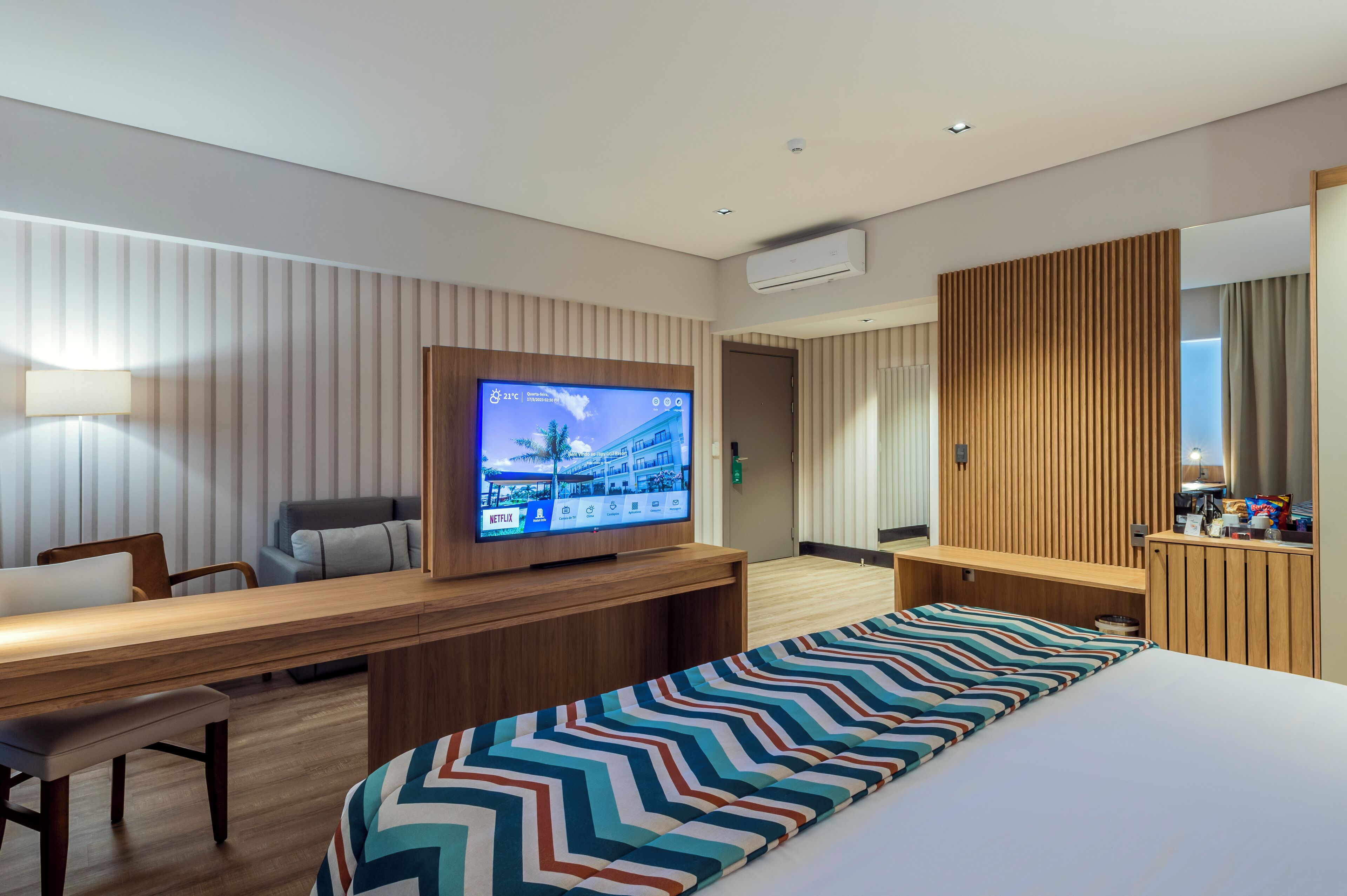 A modern hotel room with a large flat-screen TV, a wooden desk, a comfortable sofa, a bed with a colorful striped bedspread, and a wooden panel wall.
