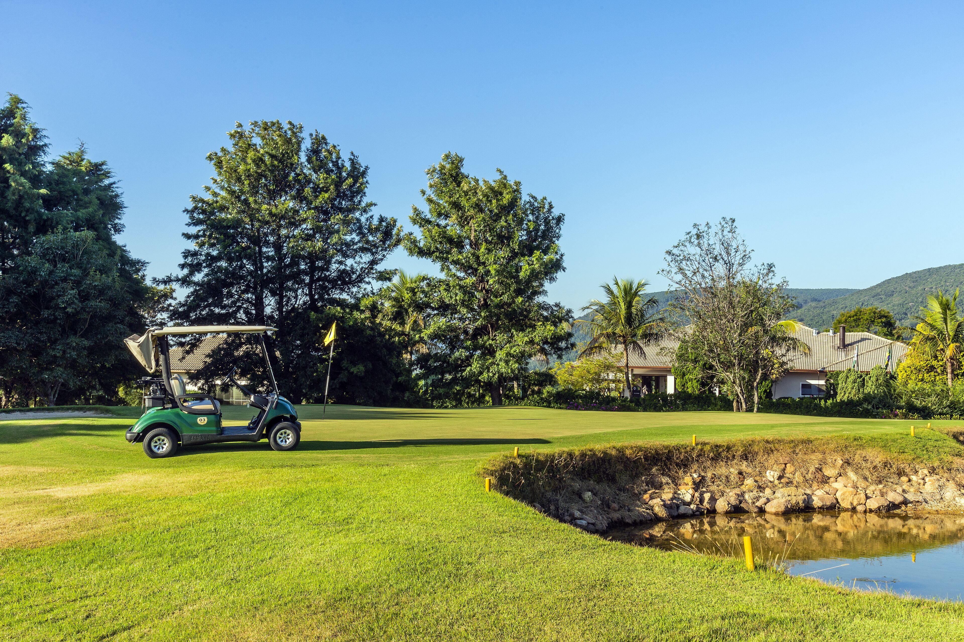 A golf course with a green cart, a pond, and a house in the background.