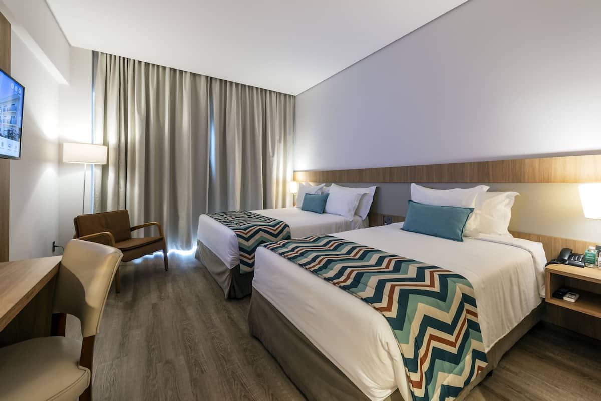A hotel room with two beds, a desk, a chair, a TV, and a nightstand with a phone and a bottle.