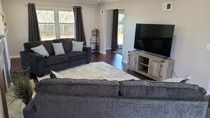 Una sala de estar moderna con un sofá gris, un televisor de pantalla plana sobre un soporte de madera y un ventanal con cortinas.