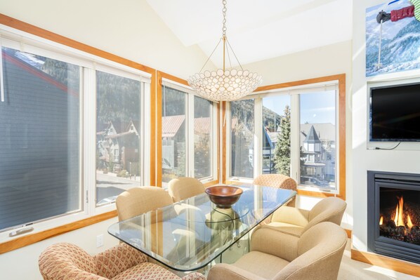 A modern living room with a glass-top dining table, beige sofas, large windows, and a fireplace.