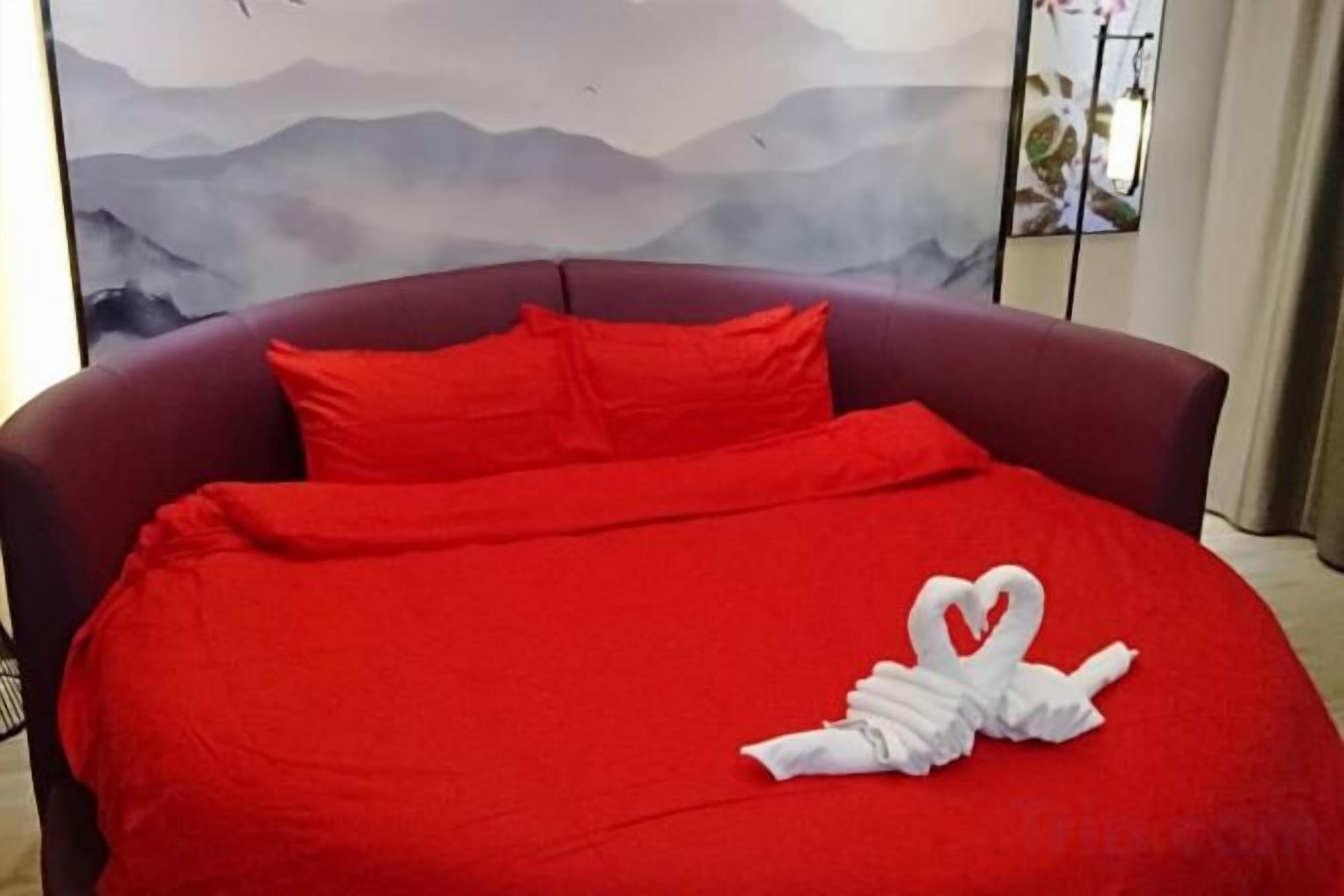 A bed with a red bedspread and pillows, a heart-shaped towel arrangement, and a painting with mountains in the background.