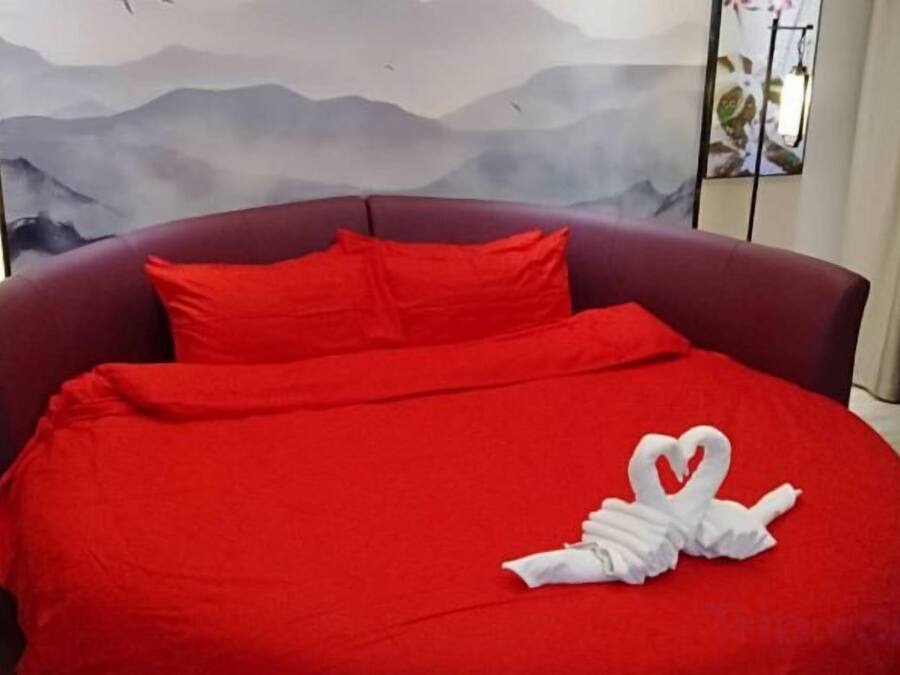 A bed with a red bedspread and pillows, a heart-shaped towel arrangement, and a painting with mountains in the background.