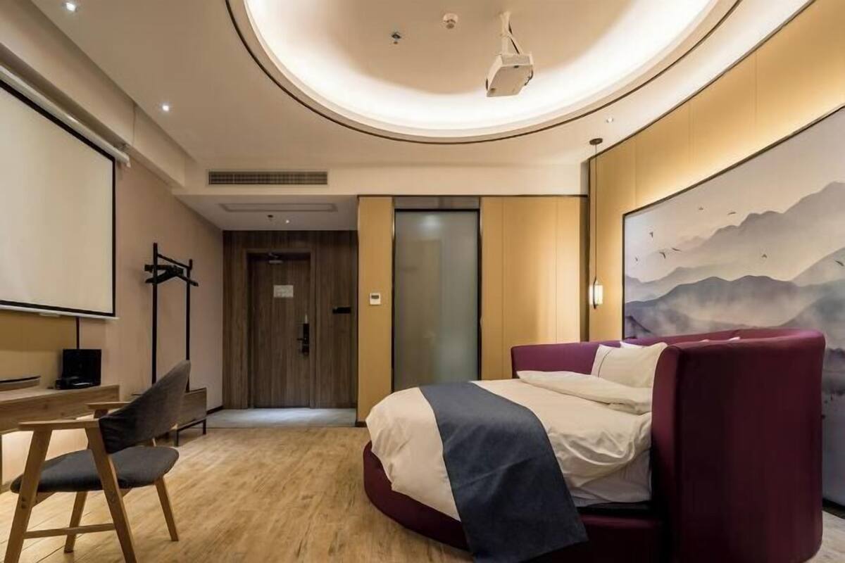 A modern hotel room with a bed, a desk, a chair, a projector screen, and a large painting on the wall.