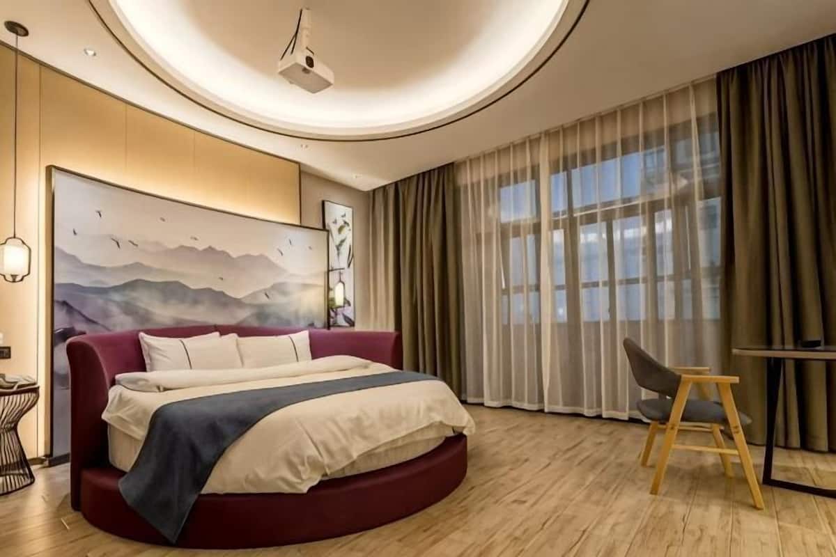 A modern hotel room with a curved bed, a desk, and a large window with curtains.