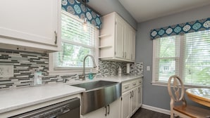 A modern kitchen with white cabinets, a stainless steel sink, and a dishwasher. There is a window with blinds and a patterned curtain. - Central Tosa home near downtown, state fair, froedtert (Wauwatosa)