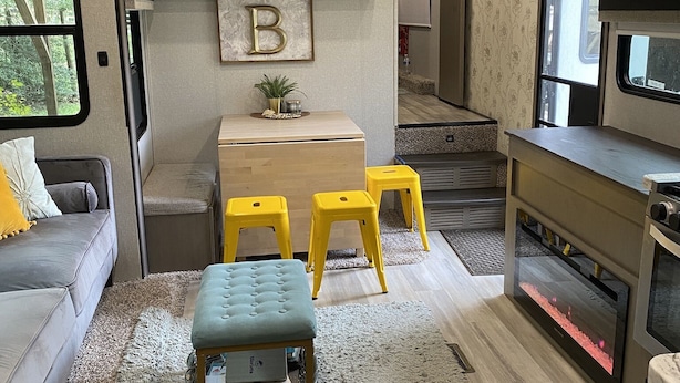 Interior compacto de autocaravana con kitchenette, zona de comedor con taburetes amarillos, sofá gris y una pequeña mesa.