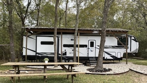 Una caravana blanca con un porche cubierto, una mesa de picnic y árboles al fondo.