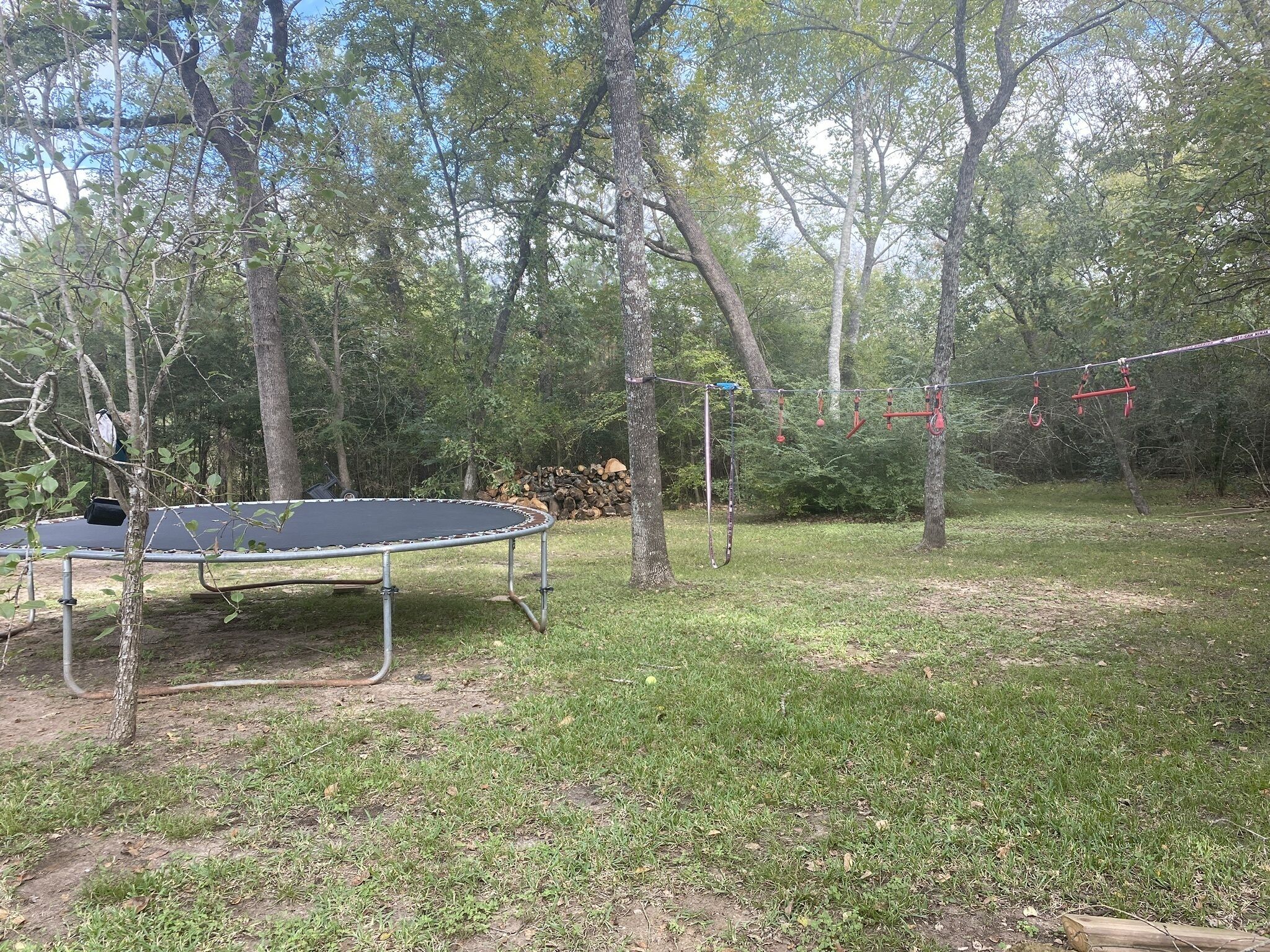 A trampoline set up in a wooded area with trees and a pile of logs in the background.
