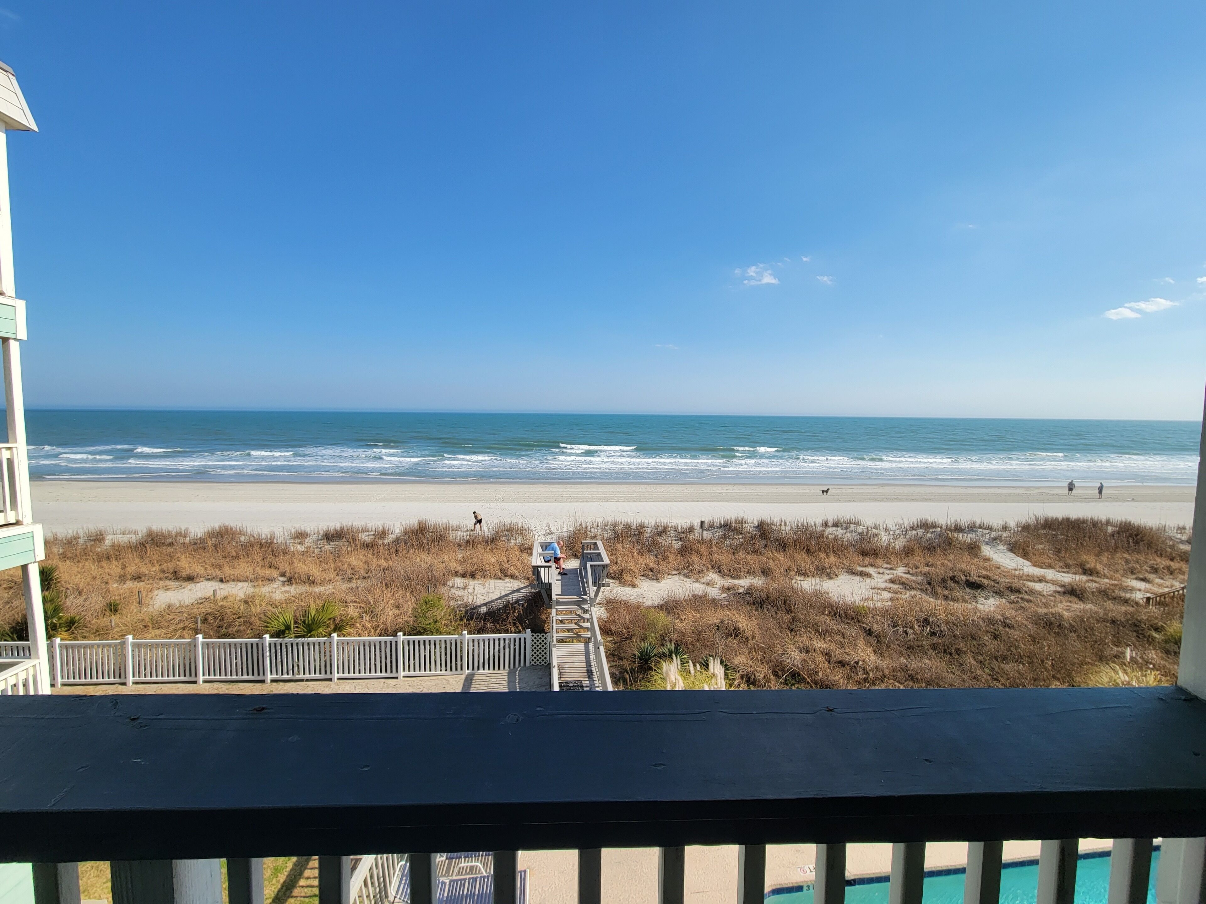 A beach view from a balcony with a wooden deck, a white fence, and a clear sky.