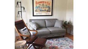 Ein Wohnzimmer mit grauem Sofa, einem Holzsesseln, einem gemusterten Teppich und einem Bilderrahmen mit einem Foto einer Pianofirma an der Wand.