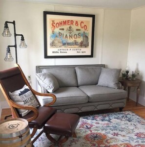 A living room with a grey sofa, a wooden armchair, a patterned rug, and a framed picture of a piano company on the wall.