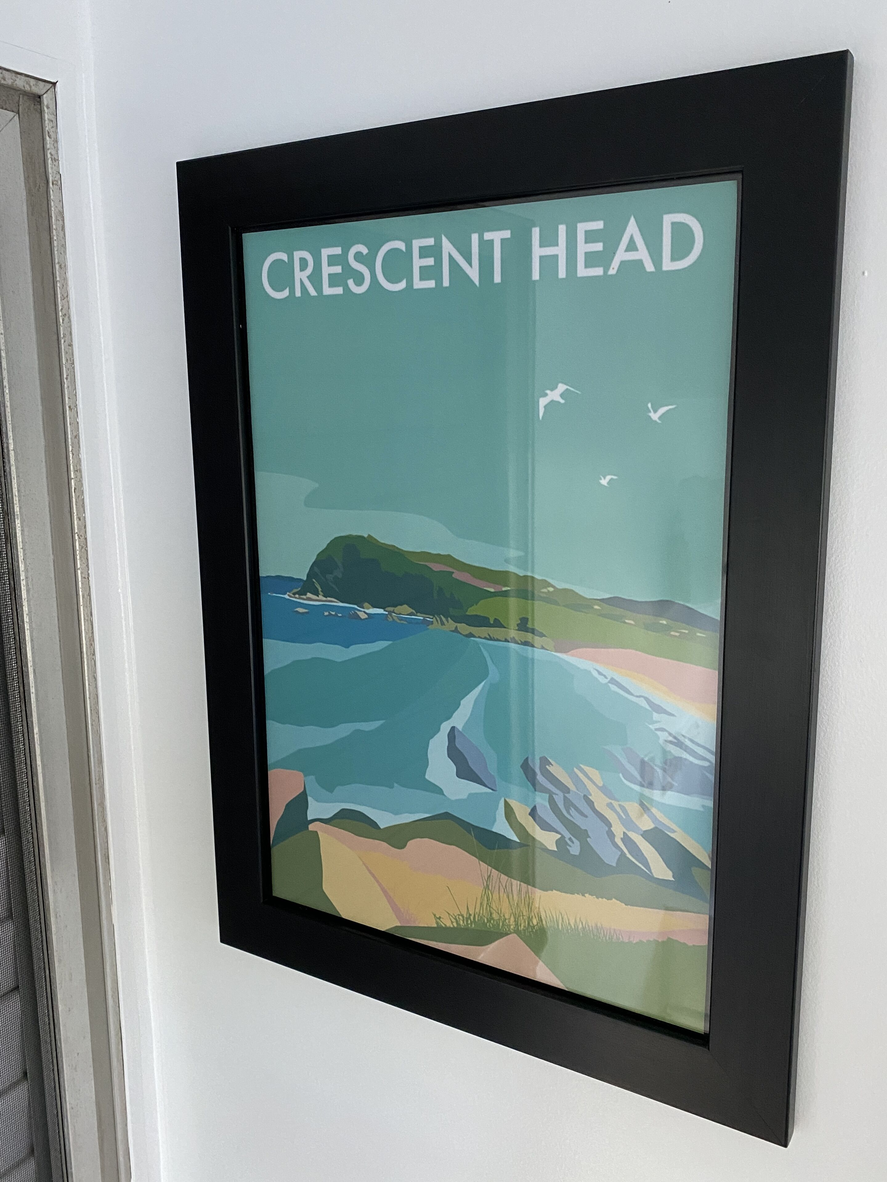 A framed artwork titled 'Crescent Head' depicting a coastal landscape with mountains and birds.