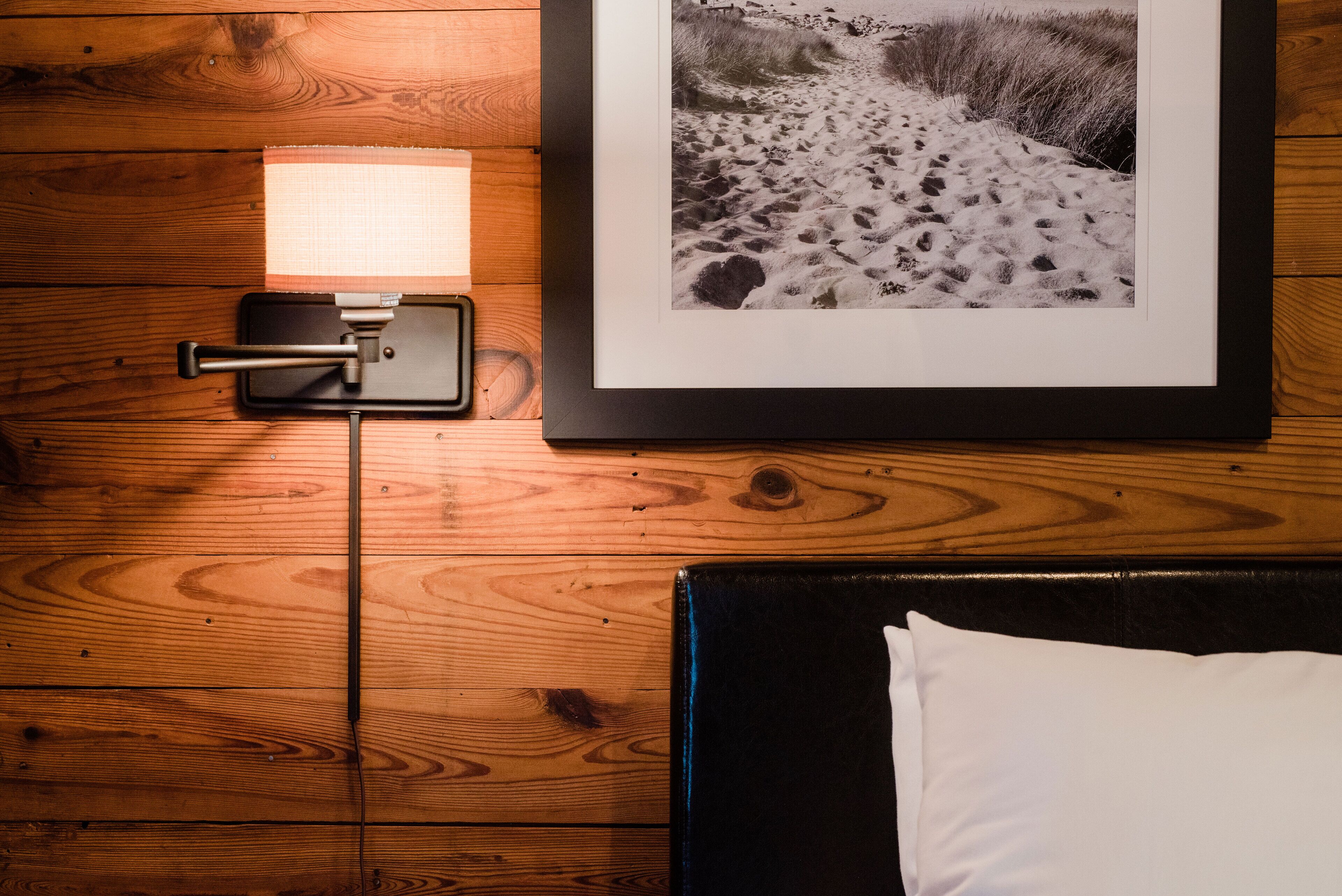 Fotografía enmarco de un sendero arenoso en la playa, una lámpara fijada a la pared y una cama con un almohadón blanco.