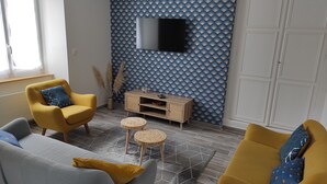 Ein Wohnzimmer mit einer Couch, Sesseln, einem Couchtisch, einem an einer gemusterten Wand montierten Fernseher und einem Schrank.