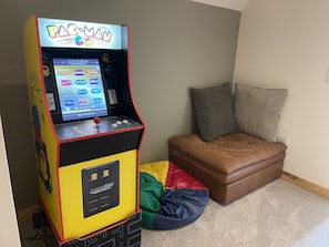 Une borne d’arcade Pac-Man, placée dans un coin d’une pièce, avec un canapé en cuir marron et un petit fauteuil en forme de sac de haricots.