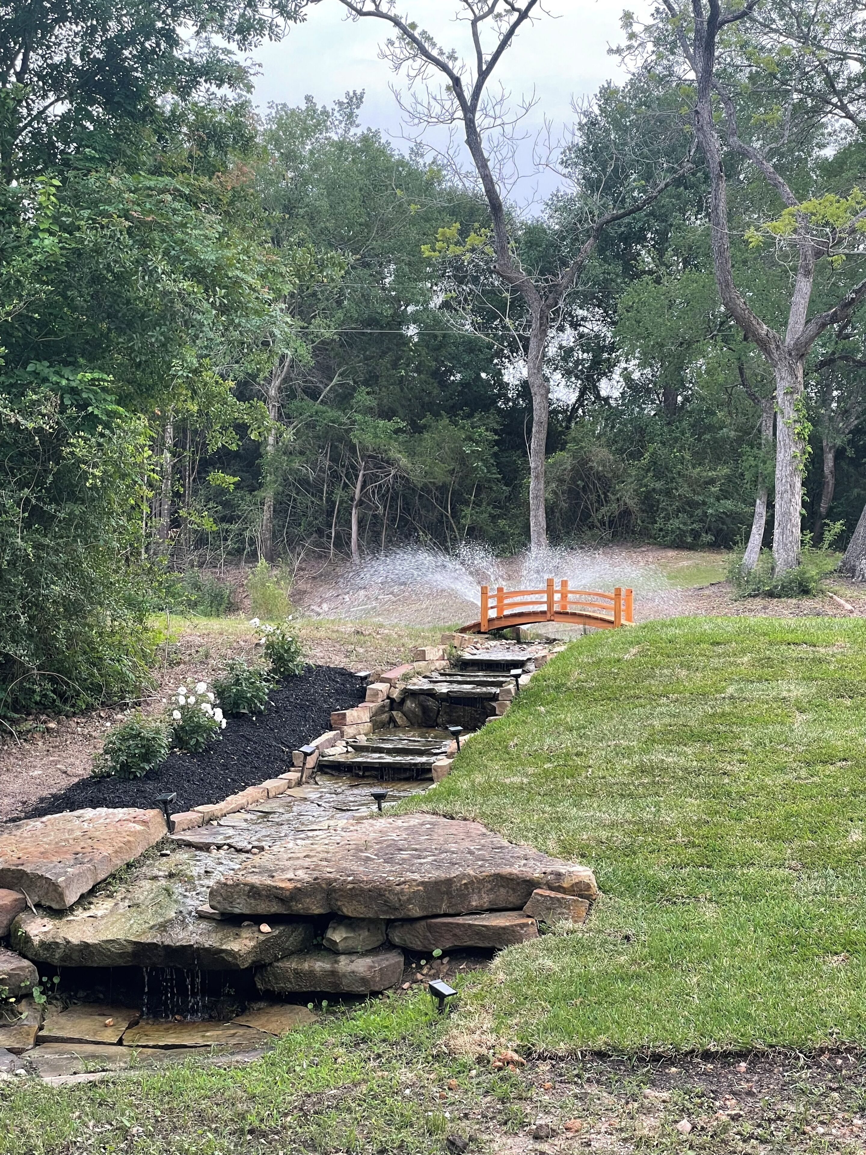 Ein Steinelement mit Wasserspeil und einer Holzbrücke in einem bewaldeten Gebiet.
