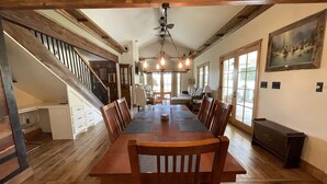 A spacious dining area with a long wooden table and chairs, a staircase on the left, and a large window on the right.