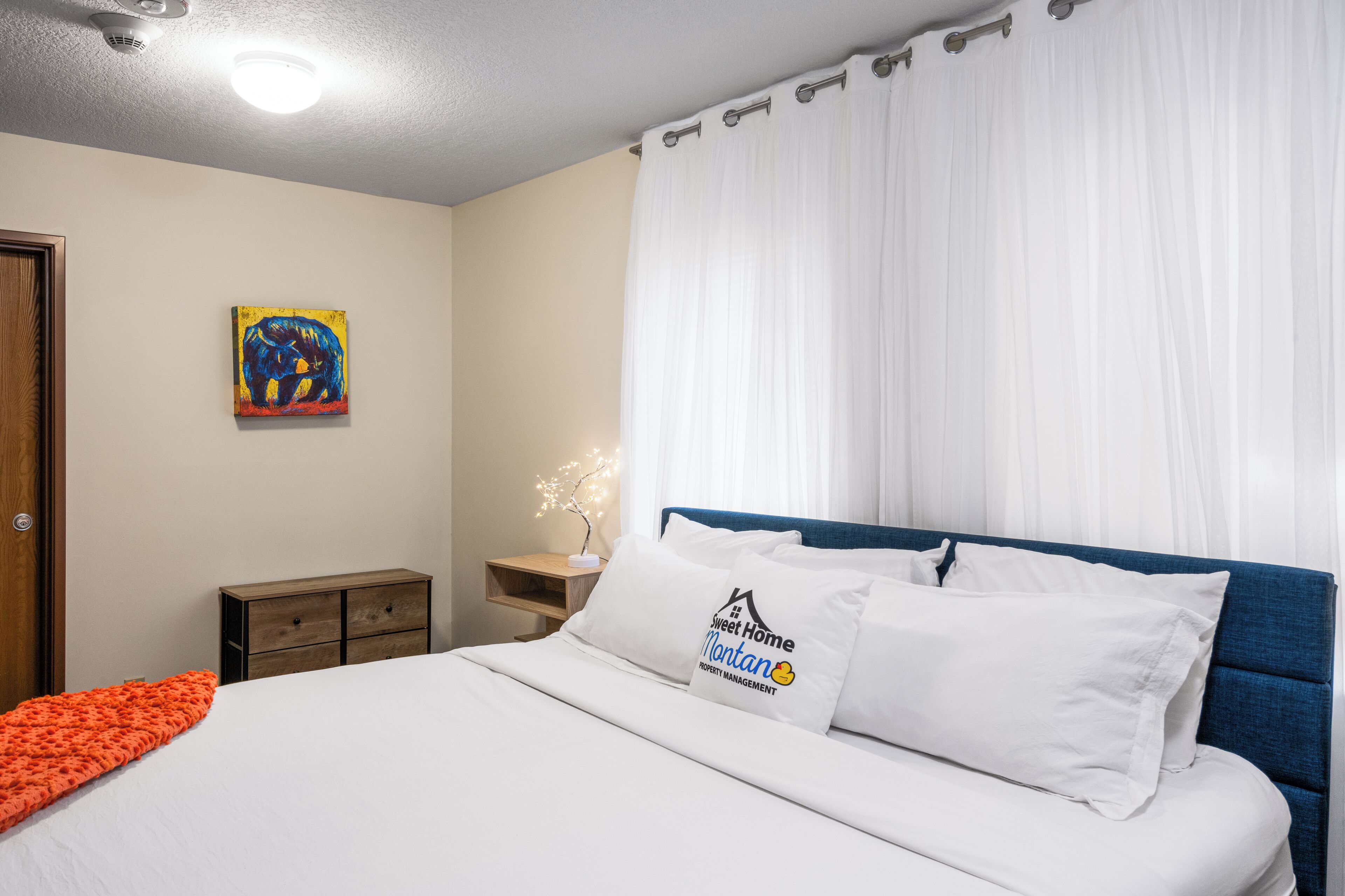 A bedroom with a bed, white bedding, a blue headboard, a nightstand with a lamp, a painting on the wall, and a window with white curtains.