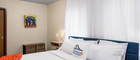 A bedroom with a bed, white bedding, a blue headboard, a nightstand with a lamp, a painting on the wall, and a window with white curtains.