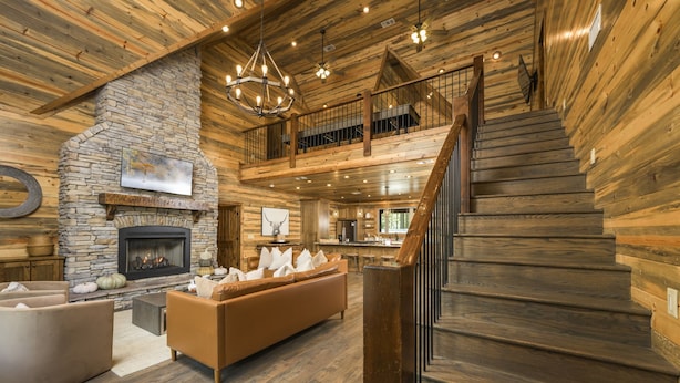 A spacious living area with a stone fireplace, a large sofa, and a wooden staircase leading to an upper level. - Luxury Forest Escape with Charm - Hot Tub, Game Loft, Pool Table and Playground (Broken Bow)
