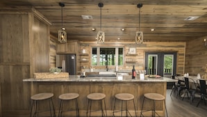 A modern kitchen with wooden cabinetry, a central island with stools, pendant lighting, and a view of the outdoors through large windows. - Luxury Forest Escape with Charm - Hot Tub, Game Loft, Pool Table and Playground (Broken Bow)