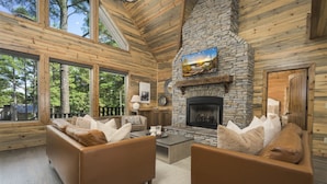 A log cabin interior with a stone fireplace, leather sofas, and large windows offering a view of the surrounding forest. - Luxury Forest Escape with Charm - Hot Tub, Game Loft, Pool Table and Playground (Broken Bow)
