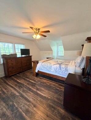 A bedroom with a bed, dresser, television, ceiling fan, and windows.